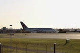 A large airplane with Qatar Airways branding is positioned on an airport runway during daylight. The aircraft is seen from a side view, and there is a control tower visible on the left side of the image. The area surrounding the runway is grassy, and the scene conveys a calm atmosphere.