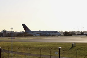 A large airplane with Qatar Airways branding is positioned on an airport runway during daylight. The aircraft is seen from a side view, and there is a control tower visible on the left side of the image. The area surrounding the runway is grassy, and the scene conveys a calm atmosphere.