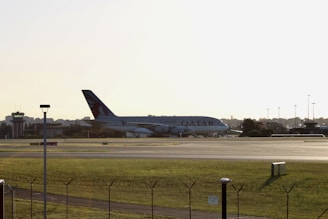 A large airplane with Qatar Airways branding is positioned on an airport runway during daylight. The aircraft is seen from a side view, and there is a control tower visible on the left side of the image. The area surrounding the runway is grassy, and the scene conveys a calm atmosphere.