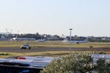 An airport scene features commercial airplanes on a runway, with one plane visibly taking off or landing, emitting a cloud of vapor or smoke. The background includes airport facilities and a large IKEA store. A security or ground vehicle is visible on a taxiway, and the foreground contains what appears to be the tops of vehicles, possibly parked cars or buses.