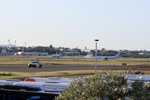 An airport scene features commercial airplanes on a runway, with one plane visibly taking off or landing, emitting a cloud of vapor or smoke. The background includes airport facilities and a large IKEA store. A security or ground vehicle is visible on a taxiway, and the foreground contains what appears to be the tops of vehicles, possibly parked cars or buses.