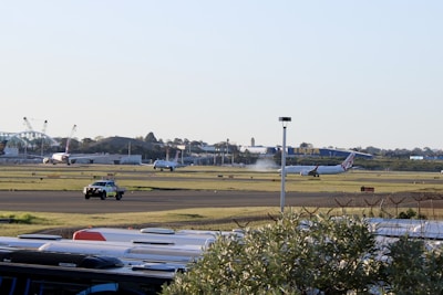 An airport scene features commercial airplanes on a runway, with one plane visibly taking off or landing, emitting a cloud of vapor or smoke. The background includes airport facilities and a large IKEA store. A security or ground vehicle is visible on a taxiway, and the foreground contains what appears to be the tops of vehicles, possibly parked cars or buses.
