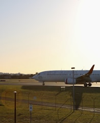 A commercial airplane bearing the livery of a well-known airline is visible on an airport runway. The aircraft is taxiing or preparing for takeoff, captured during golden hour with a warm sunlight casting long shadows. A fence with a parking sign in the foreground and airport infrastructure are visible in the distance.
