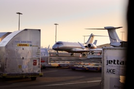 An aircraft is parked on an airport tarmac surrounded by storage containers labeled with logistics company brands. The scene is illuminated by a soft, warm light from the setting or rising sun, and several airport light poles are visible in the background.