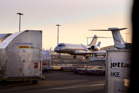 An aircraft is parked on an airport tarmac surrounded by storage containers labeled with logistics company brands. The scene is illuminated by a soft, warm light from the setting or rising sun, and several airport light poles are visible in the background.