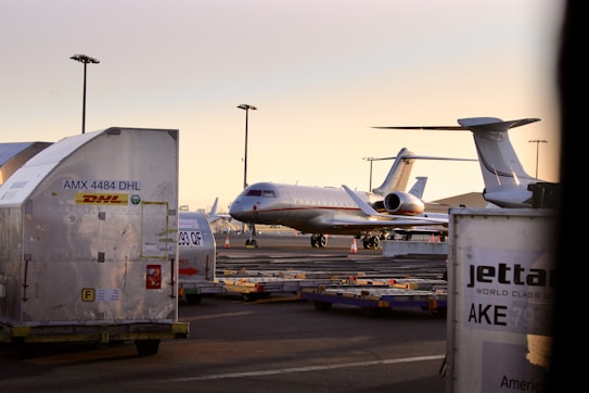 An aircraft is parked on an airport tarmac surrounded by storage containers labeled with logistics company brands. The scene is illuminated by a soft, warm light from the setting or rising sun, and several airport light poles are visible in the background.