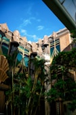 An ornate building facade features intricate architectural details with art deco style elements, including stylized geometric patterns and fluted designs. Tall, lush green palm trees partially obscure the structure, adding a tropical element to the scene. The sky is clear and blue, complementing the building's golden and earthy tones.