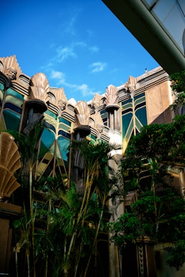 An ornate building facade features intricate architectural details with art deco style elements, including stylized geometric patterns and fluted designs. Tall, lush green palm trees partially obscure the structure, adding a tropical element to the scene. The sky is clear and blue, complementing the building's golden and earthy tones.