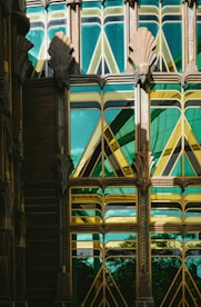 An intricate Art Deco architectural facade featuring geometric patterns and stylized motifs. The building uses a combination of green and gold reflective glass, framed by ornate metalwork. The design includes symmetrical fan-like decorations and angular shapes, creating a visually striking and harmonious appearance.