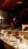 A chef is preparing food behind a wooden counter in a dimly lit restaurant. The counter is set with a variety of plates, cups, and utensils. In the background, decorative elements such as a menu board with writing and shelves with bottles contribute to the ambiance.