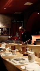 A chef is preparing food behind a wooden counter in a dimly lit restaurant. The counter is set with a variety of plates, cups, and utensils. In the background, decorative elements such as a menu board with writing and shelves with bottles contribute to the ambiance.