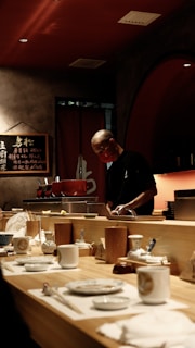 A chef is preparing food behind a wooden counter in a dimly lit restaurant. The counter is set with a variety of plates, cups, and utensils. In the background, decorative elements such as a menu board with writing and shelves with bottles contribute to the ambiance.