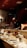 A chef is preparing food behind a wooden counter in a dimly lit restaurant. The counter is set with a variety of plates, cups, and utensils. In the background, decorative elements such as a menu board with writing and shelves with bottles contribute to the ambiance.