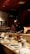 A chef is preparing food behind a wooden counter in a dimly lit restaurant. The counter is set with a variety of plates, cups, and utensils. In the background, decorative elements such as a menu board with writing and shelves with bottles contribute to the ambiance.