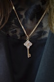 A gold chain necklace with a pendant in the shape of a key, embellished with clear, sparkling gemstones. The pendant hangs against a black, lacy textured fabric, which adds an elegant contrast to the jewelry.