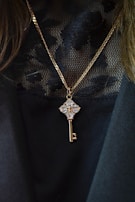 A gold chain necklace with a pendant in the shape of a key, embellished with clear, sparkling gemstones. The pendant hangs against a black, lacy textured fabric, which adds an elegant contrast to the jewelry.