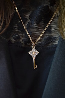 A gold chain necklace with a pendant in the shape of a key, embellished with clear, sparkling gemstones. The pendant hangs against a black, lacy textured fabric, which adds an elegant contrast to the jewelry.