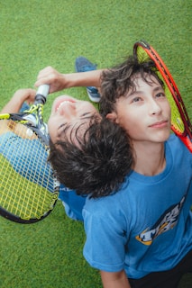 Two young individuals are seated on a grassy surface, leaning against each other with their heads together. They are holding tennis rackets with vibrant color patterns. One person is smiling broadly, while the other looks thoughtful. The setting appears joyful and relaxed.
