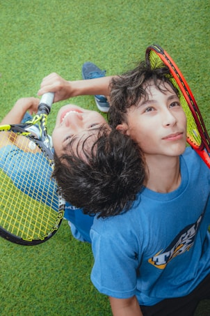 Two young individuals are seated on a grassy surface, leaning against each other with their heads together. They are holding tennis rackets with vibrant color patterns. One person is smiling broadly, while the other looks thoughtful. The setting appears joyful and relaxed.