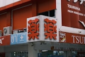 A storefront with Japanese and English signage. The main sign features large red characters on a white background, indicating a pharmacy. Additional signs include symbols for medical services and text in English and Japanese. Air conditioning units are visible near the top left.