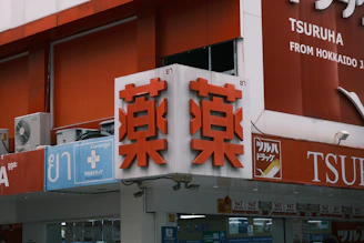A storefront with Japanese and English signage. The main sign features large red characters on a white background, indicating a pharmacy. Additional signs include symbols for medical services and text in English and Japanese. Air conditioning units are visible near the top left.
