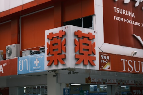A storefront with Japanese and English signage. The main sign features large red characters on a white background, indicating a pharmacy. Additional signs include symbols for medical services and text in English and Japanese. Air conditioning units are visible near the top left.