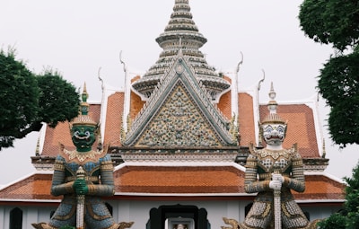 Two ornate guardian statues stand at the entrance of a Thai temple. The temple features intricate architectural details with ornate patterns, a central spire, and a tiled roof. The statues are elaborately decorated with colorful patterns and hold weapons, representing protection. Lush green trees frame the scene.