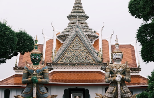 Two ornate guardian statues stand at the entrance of a Thai temple. The temple features intricate architectural details with ornate patterns, a central spire, and a tiled roof. The statues are elaborately decorated with colorful patterns and hold weapons, representing protection. Lush green trees frame the scene.