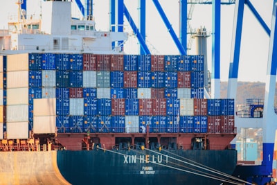 A large cargo ship is docked at a port with numerous stacked shipping containers in various colors such as blue, red, and white. The ship has cranes towering above it and appears to be engaged in loading or unloading operations.