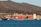 A large cargo ship loaded with colorful shipping containers is docked at a port, set against a backdrop of rolling hills and industrial buildings. The ship is painted blue with a white superstructure, and the containers are stacked in orderly rows. The surrounding water is calm, with a deep blue hue.