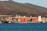 A large cargo ship loaded with colorful shipping containers is docked at a port, set against a backdrop of rolling hills and industrial buildings. The ship is painted blue with a white superstructure, and the containers are stacked in orderly rows. The surrounding water is calm, with a deep blue hue.