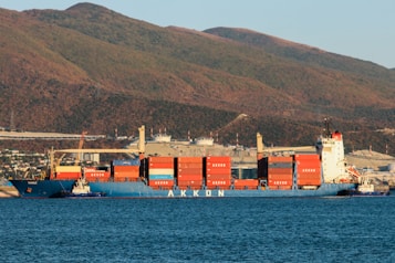 A large cargo ship loaded with colorful shipping containers is docked at a port, set against a backdrop of rolling hills and industrial buildings. The ship is painted blue with a white superstructure, and the containers are stacked in orderly rows. The surrounding water is calm, with a deep blue hue.