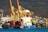 Large cranes with yellow and red structures dominate the port area. A container ship named 'NARA ISTANBUL' is docked, loaded with numerous colorful shipping containers. In the background, a hillside is dotted with small buildings, contrasting against the industrial scene.