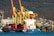 Large cranes with yellow and red structures dominate the port area. A container ship named 'NARA ISTANBUL' is docked, loaded with numerous colorful shipping containers. In the background, a hillside is dotted with small buildings, contrasting against the industrial scene.
