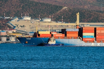 A large cargo ship loaded with numerous colorful containers is situated in the harbor. The ship is accompanied by a smaller tugboat that is positioned at its side. Behind the vessels, there is an industrial area with storage tanks, buildings, and infrastructure extending up a hillside. The clear blue water of the harbor contrasts with the backdrop of hills covered with greenery and a partially overcast sky.