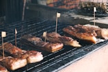 Several pieces of grilled or barbecued meat laid on a grid iron grill, each piece is marked with a small flag-like skewer. The meat has a crispy, charred appearance with visible caramelization, and there is a rustic setting with a brick wall in the background.