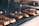 Several pieces of grilled or barbecued meat laid on a grid iron grill, each piece is marked with a small flag-like skewer. The meat has a crispy, charred appearance with visible caramelization, and there is a rustic setting with a brick wall in the background.