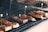Several pieces of grilled or barbecued meat laid on a grid iron grill, each piece is marked with a small flag-like skewer. The meat has a crispy, charred appearance with visible caramelization, and there is a rustic setting with a brick wall in the background.