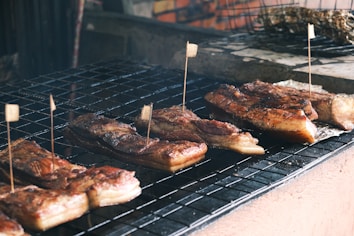 Several pieces of grilled or barbecued meat laid on a grid iron grill, each piece is marked with a small flag-like skewer. The meat has a crispy, charred appearance with visible caramelization, and there is a rustic setting with a brick wall in the background.