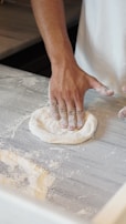 A baker sprinkling flour over dough on a floured countertop.