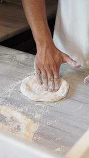 A baker sprinkling flour over dough on a floured countertop.