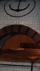 A pizza oven with a tiled mosaic design featuring an anchor symbol. Inside the oven, there is a pizza baking on the stone surface. The oven has a rustic appearance with a paddle handle slightly visible in the foreground.