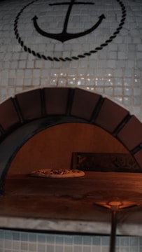 A pizza oven with a tiled mosaic design featuring an anchor symbol. Inside the oven, there is a pizza baking on the stone surface. The oven has a rustic appearance with a paddle handle slightly visible in the foreground.