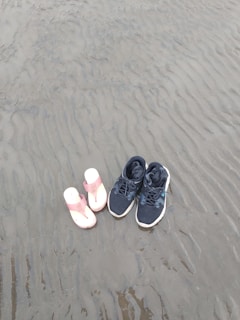 Two pairs of footwear are placed on wet, rippled sand. On the left is a pair of pink and white sandals, and on the right is a pair of black athletic shoes. The sand shows patterns formed by the receding tide, creating a natural, textured background.