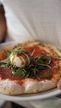 A close-up of a pizza with a thick, slightly charred crust. The toppings include a layer of tomato sauce, fresh arugula leaves, and a ball of creamy burrata cheese drizzled with balsamic glaze.