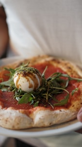 A close-up of a pizza with a thick, slightly charred crust. The toppings include a layer of tomato sauce, fresh arugula leaves, and a ball of creamy burrata cheese drizzled with balsamic glaze.