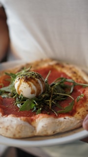 A close-up of a pizza with a thick, slightly charred crust. The toppings include a layer of tomato sauce, fresh arugula leaves, and a ball of creamy burrata cheese drizzled with balsamic glaze.