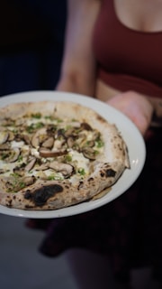 A person holding a white plate with a wood-fired pizza topped with mushrooms, herbs, and cheese. The crust appears golden brown with some charred spots.