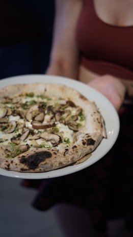 A person holding a white plate with a wood-fired pizza topped with mushrooms, herbs, and cheese. The crust appears golden brown with some charred spots.
