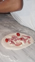 A partially prepared pizza dough topped with slices of prosciutto, cherry tomatoes, and chunks of cheese rests on a lightly floured surface. An arm wearing a white sleeve is visible in the background.
